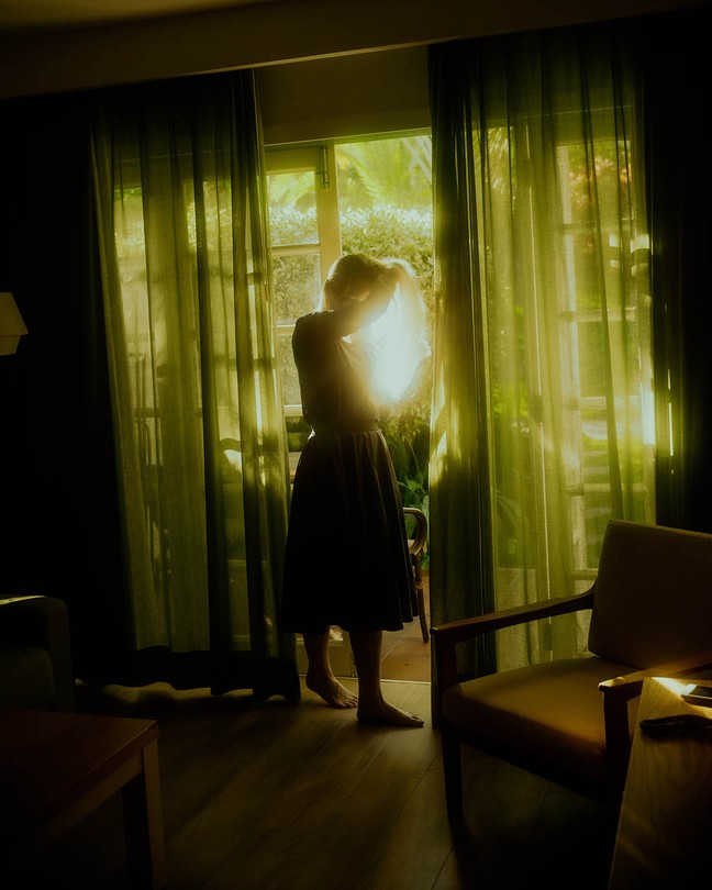 A woman in a dress peering through a sunlit window