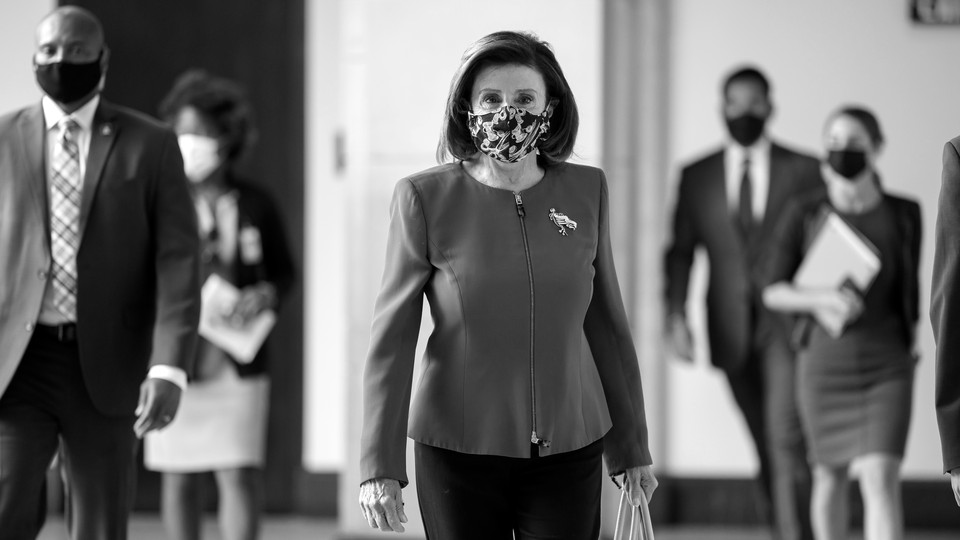 A black and white photograph of Nancy Pelosi walking toward the camera. She is wearing a pant suit and floral mask.