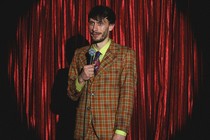 Richard Gadd in an orange plaid suit onstage, holding a microphone in front of red curtains