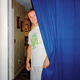 photo of man wearing white t-shirt with turtle logo and shorts looks out from behind a blue curtain inside a home