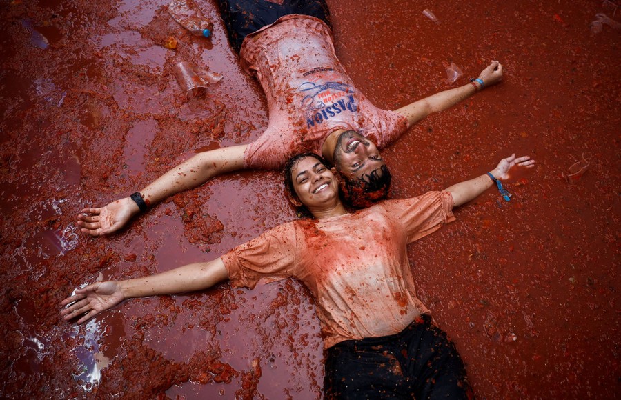 Two people lie on their backs, smiling, in a street covered in tomato pulp.