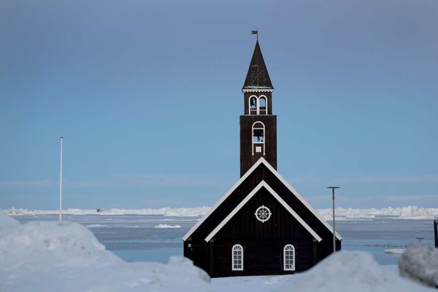 A church painted black stands on a snow-covered shoreline, with icebergs visible in the distance.