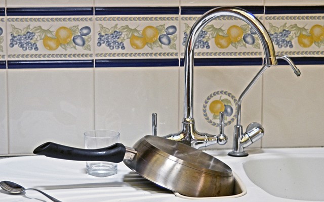 A kitchen sink with a pan, a glass, and a spoon sitting on the counter next to it.