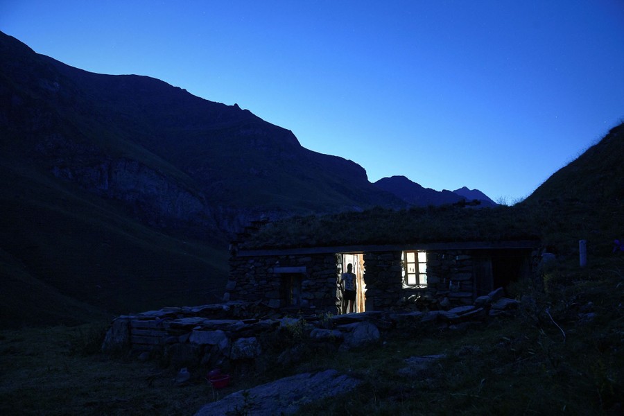 A person stands in the doorway of a small cabin in a mountain valley.