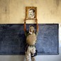 A U.S. Marine, photographed from behind, raises his hands to grip the frame of a portrait of Saddam Hussein hanging above a blackboard.
