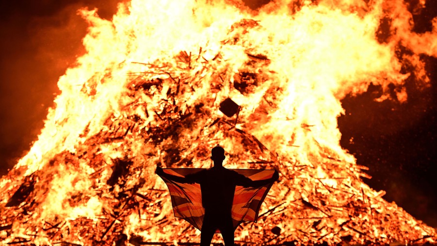 A person holding a flag stands before a huge bonfire.
