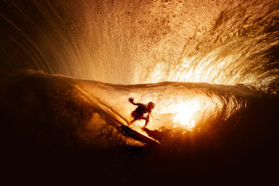 A surfer on a wave is seen in silhouette, from underwater, against the light of a sunset.