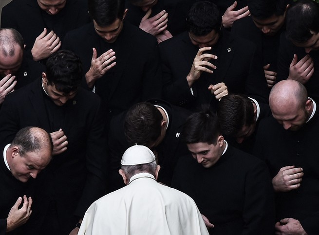 Pope Francis, with his back turned away, prays with priests.