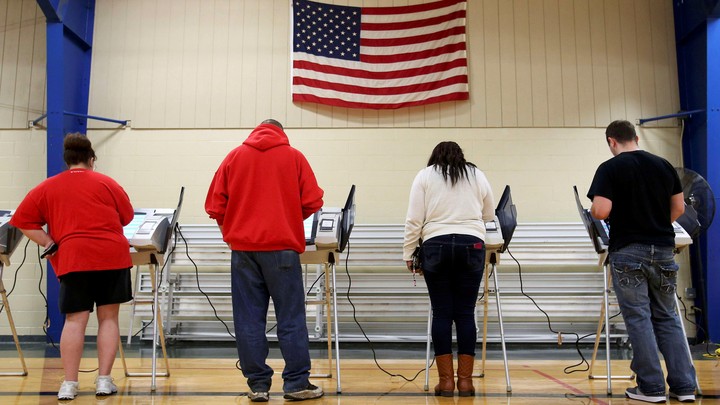 The backs of voters as they cast their ballots. They stand underneath an American flag, presumably in a gymnasium.
