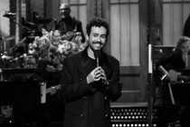Ramy Youssef smiles on the SNL stage while holding a microphone
