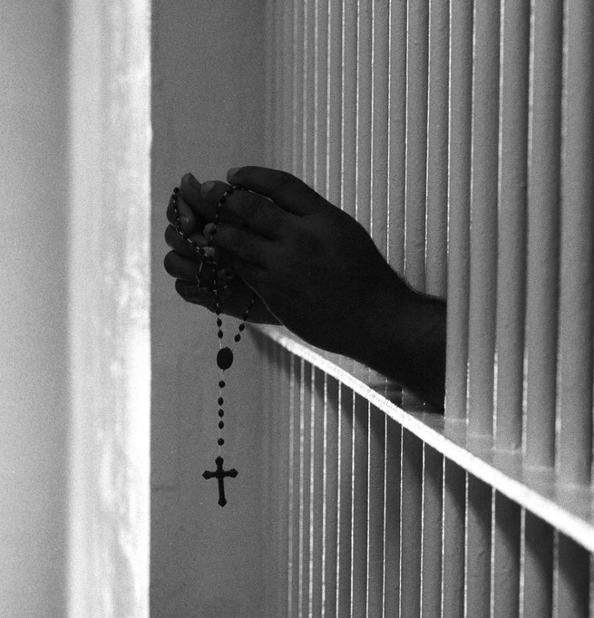 an inmates hands shown through bars holding a rosary