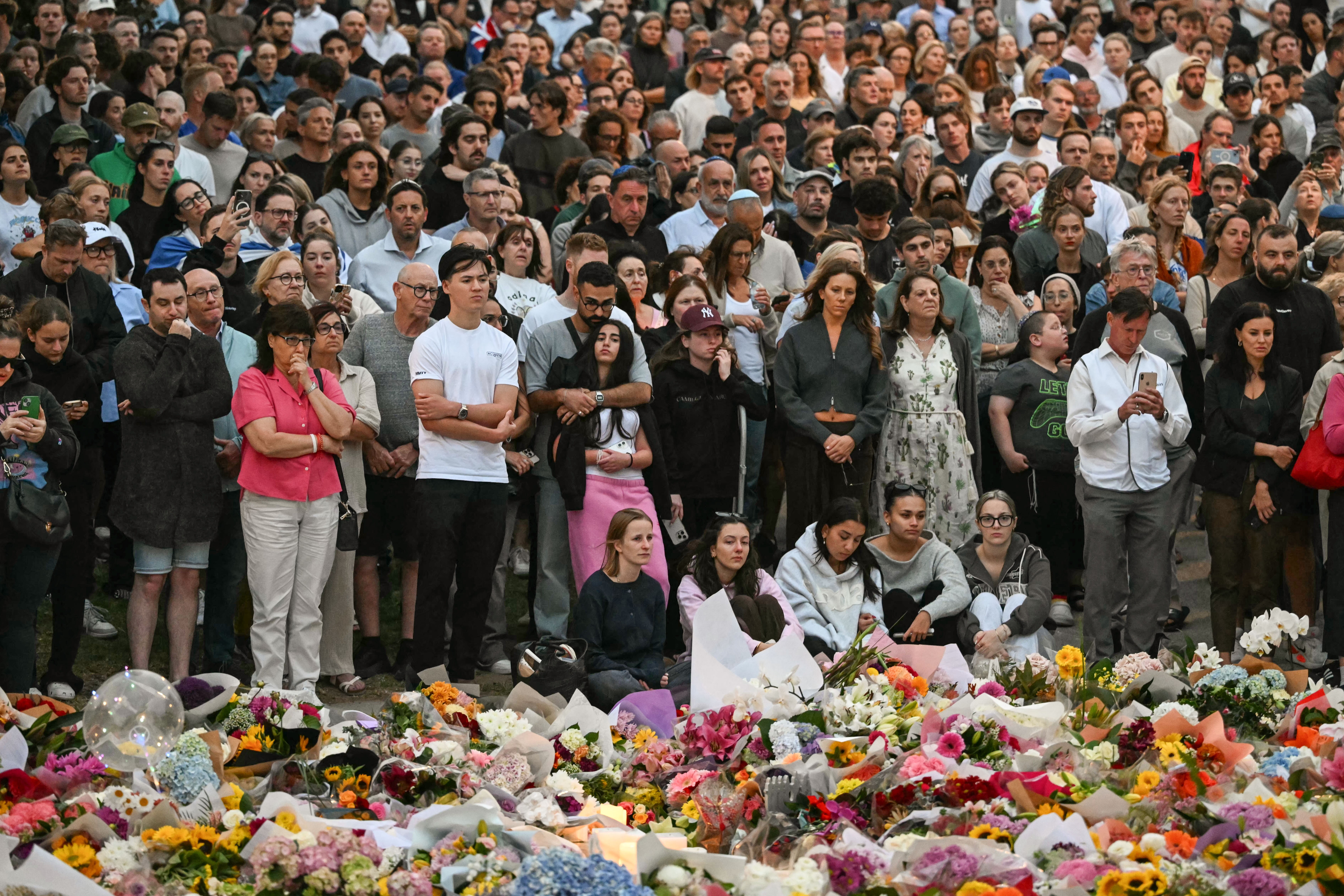 A large group of mourners gather at a tribute, standing beside piles of flower bouquets.