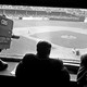 A black and white photograph of the CBS press box with one person monitoring a camera on the left, and two other figures, looking onto a baseball diamond