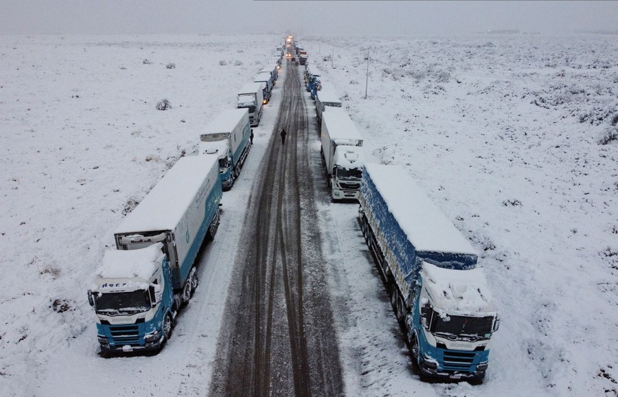 Dozens of freight trucks sit parked on either side of a snow-covered highway.