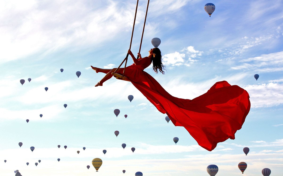 A person rides a swing wearing a flowing skirt with hot air balloons flying in the background.