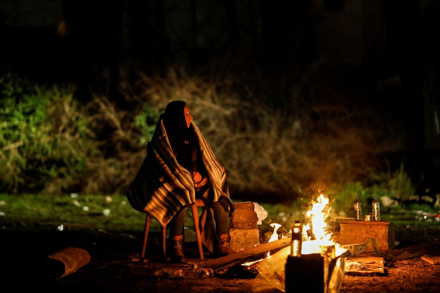 A woman wrapped in a blanket sits next to a camp fire.