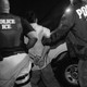 Black-and-white photograph of two ICE officers escorting a handcuffed detainee beside a white van