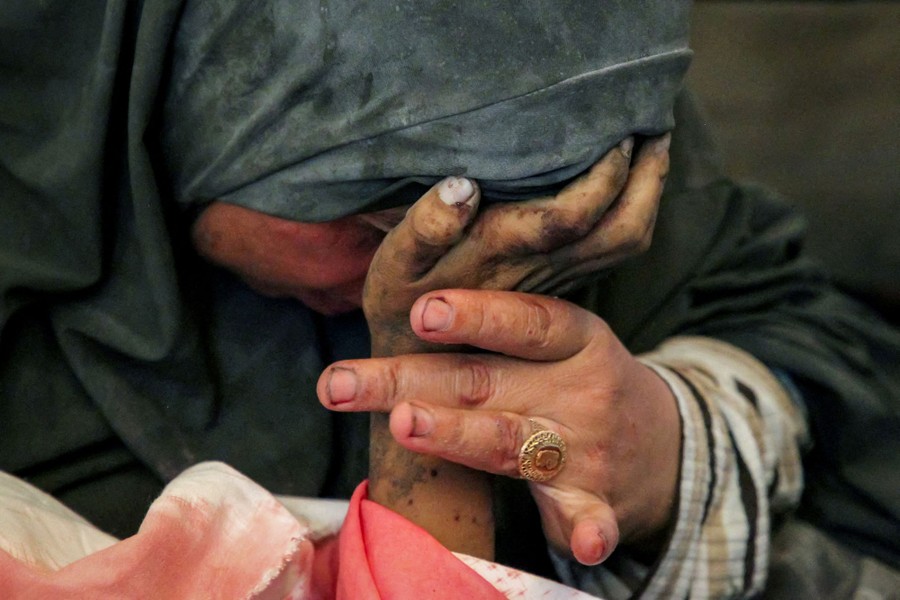 A woman leans close to the wrapped body of her child, holding his bloodied hand to her forehead.