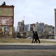 Two people walk past a vacant lot and boarded-up homes