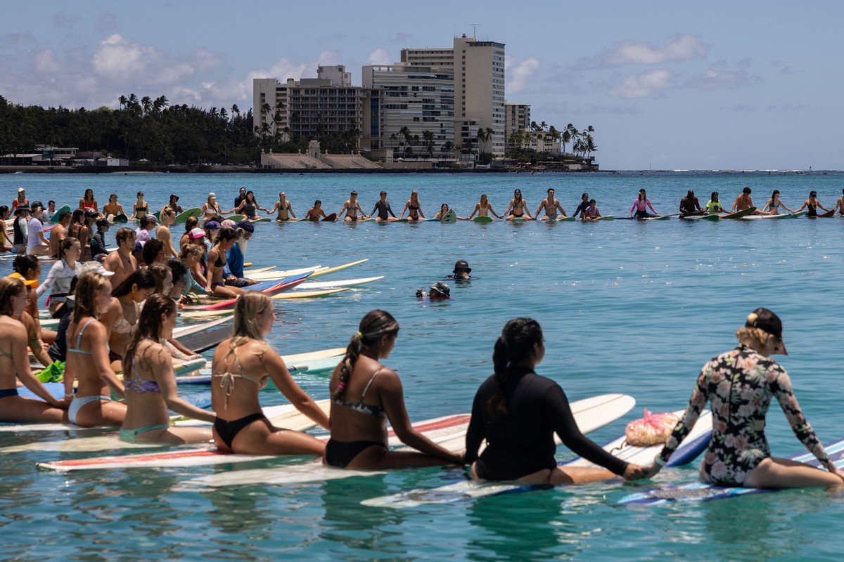 Dozens of people sit on surfboards, floating in a circle, holding hands, near a beach.