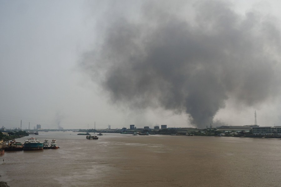 Dark smoke rises in the distance, seen among buildings on the other side of a river.