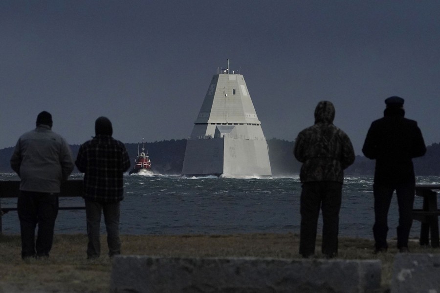 Several people watch as a modern steep-walled warship travels down a river.