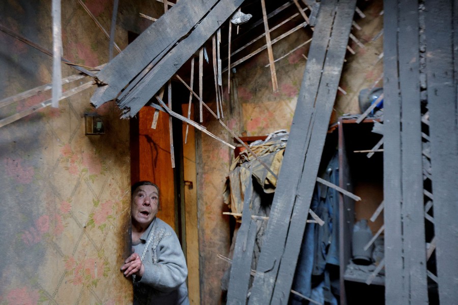 A woman leans through a doorway, looking up at the smashed ceiling of a house damaged by the fighting.
