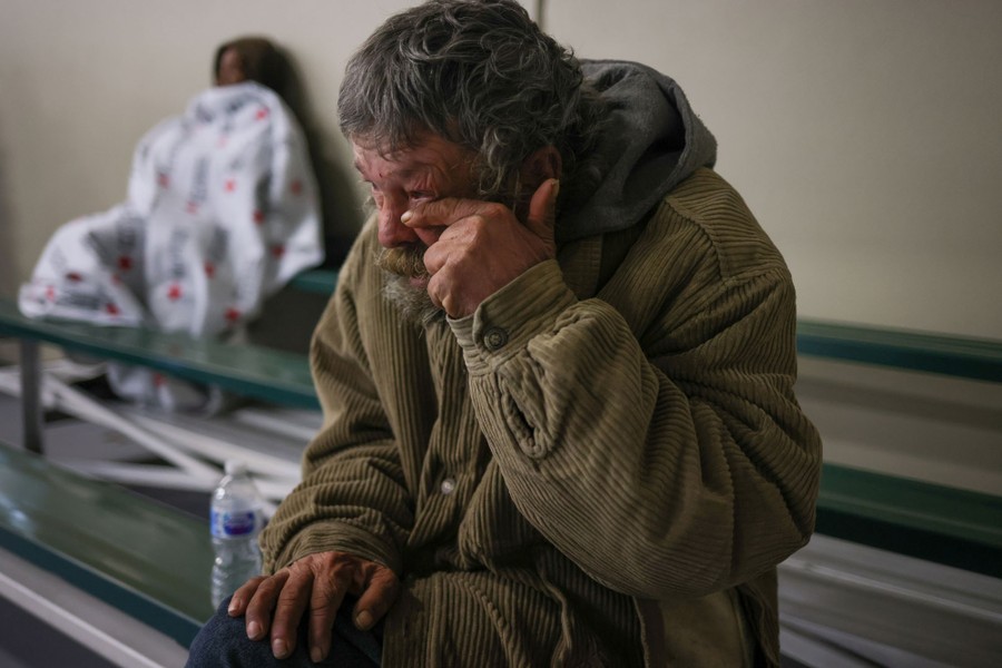 A man wipes a tear from his eye, seated on bleachers in a shelter.