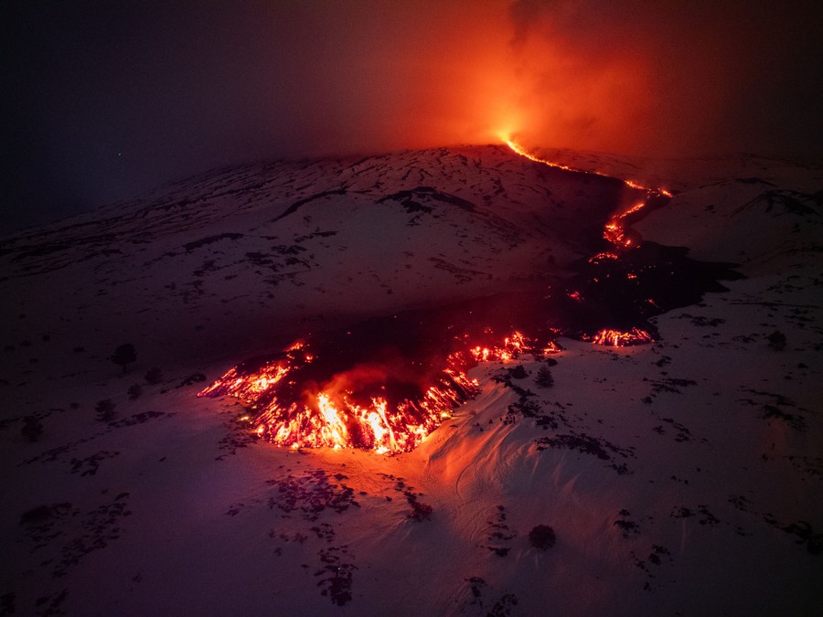 Lava flows from a fracture on Mount Etna, illuminating the surrounding snow-covered slope, at night.