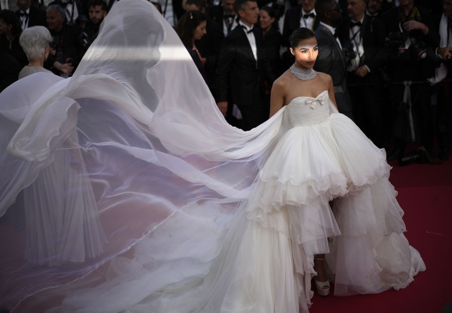 A woman poses on a red carpet, wearing a flowing white dress.