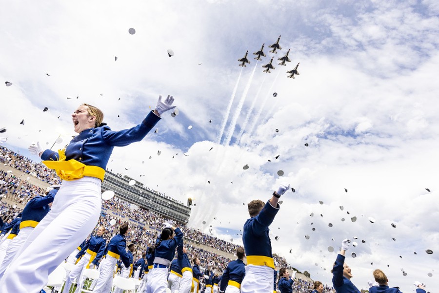 Military graduates cheer and throw their hats in the air as jets fly overhead in formation.
