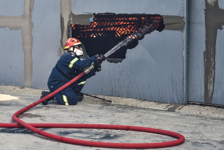 A firefighter uses a hose to spray water through a hole in a wall, into a burning building.