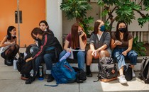 High-school students wearing masks sit together outside a school building.