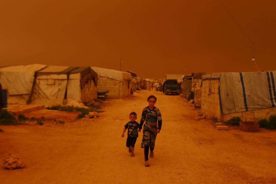 Two children walk past tents under an orange dust-filled sky.