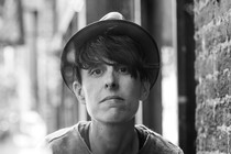 A black-and-white photo of a person with dark hair wearing a narrow hat and a thin sweater beside a brick wall