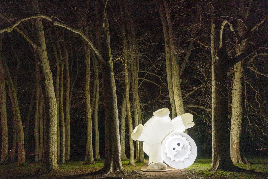 An illuminated art installation, representing a cartoonish small figure wearing thick winter clothes, set among trees