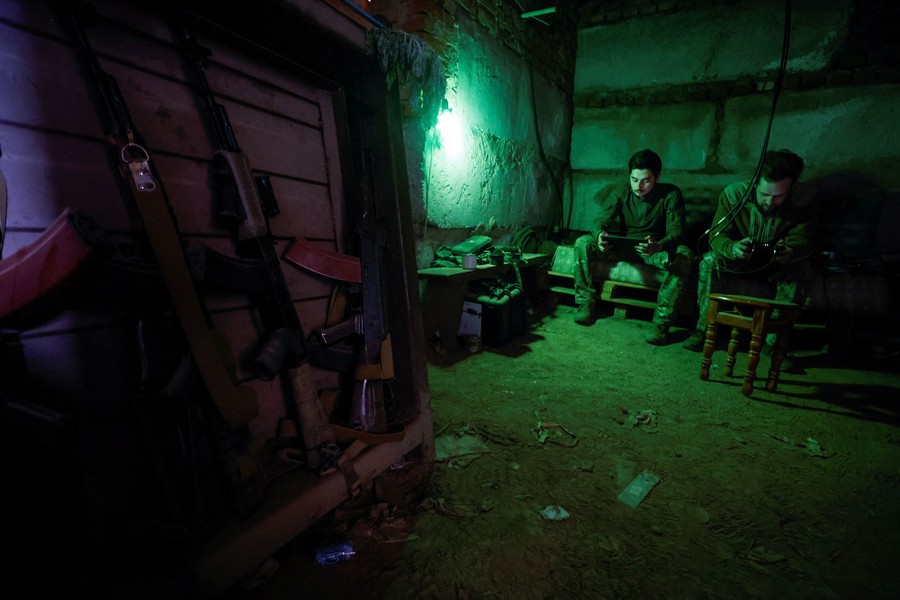 Two soldiers sit in a a dark room, holding controllers.