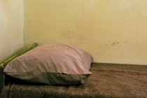 A twin bed in the corner of a room, with a thin brown blanket and a pink pillowcase, against a stained wall