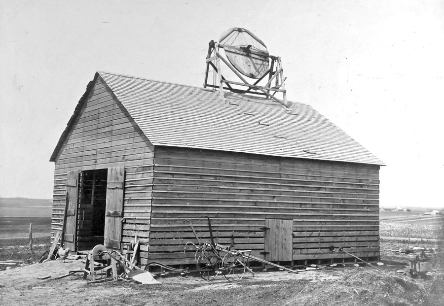 A farm shed with a circular wooden windmill mounted on its roof