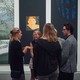 A group of five students stands, chatting, in front of three works of art: two paintings of tree branches and one portrait of a woman's face.