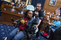 A protester in a motorized chair is pushed out of a House committee room