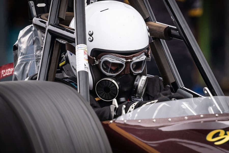 A race-car driver wearing a protective helmet and mask sits behind the steering wheel of a dragster.