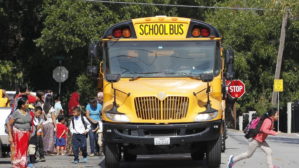 The front of a school bus fills most of the frame, as children and parents with backpacks mill about around the front of the bus. The stop sign is out on the side of the bus.