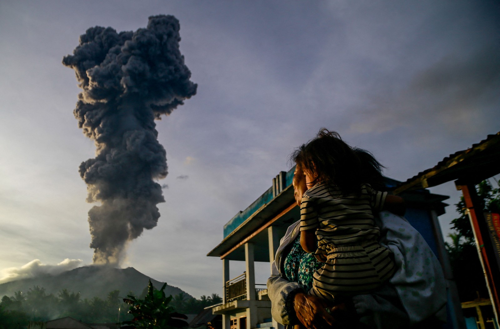 A woman and child look on at volcanic ash rising into the air.