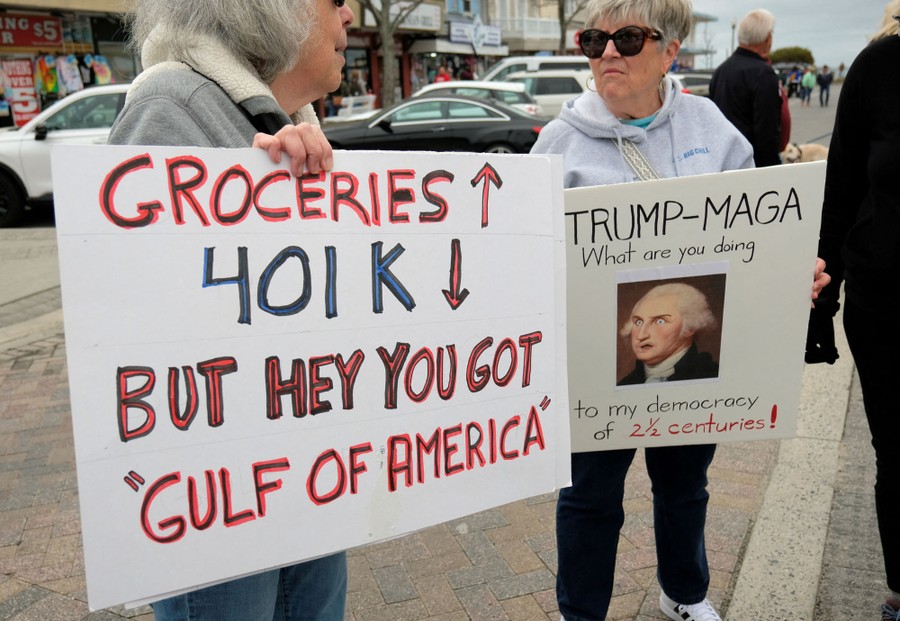 Two protesters are seen standing on a street, holding signs, including one that reads "Groceries ↑ 401k ↓ But hey you got 'Gulf of America'"