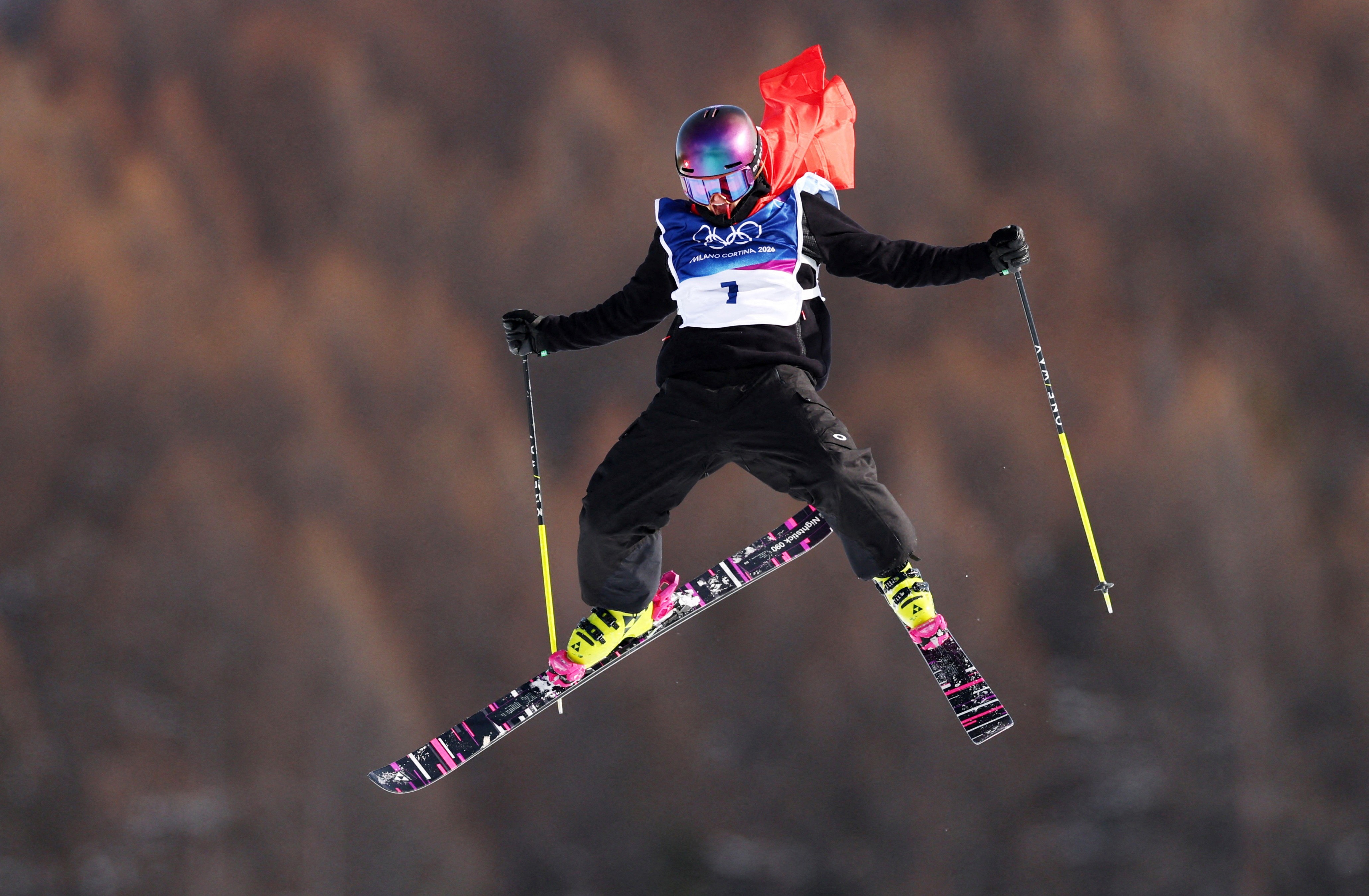 A skier, wearing a Swiss flag as a cape, celebrates while mid-jump during a ski run.