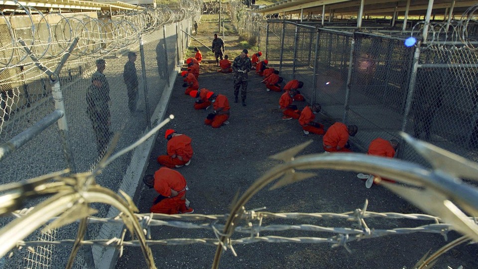 Detainees in orange jumpsuits sit in a holding area under the watchful eyes of military police during in-processing to the temporary detention facility at Camp X-Ray of Naval Base Guantanamo Bay on January 11, 2002.