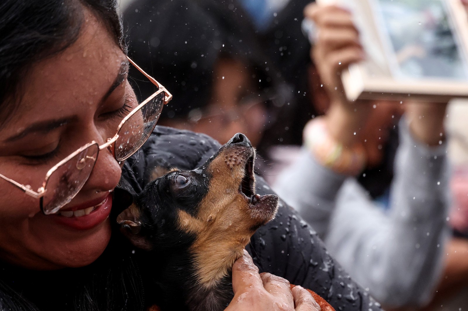 A woman holds a small dog that barks as it is sprinkled with holy water.