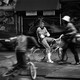 A Black teenager, in focus, sits on a bike with his arms folded, as people and cars around him are blurred.