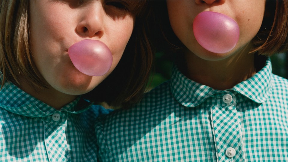 two girls wearing matching green gingham shirts and blowing pink gum bubbles